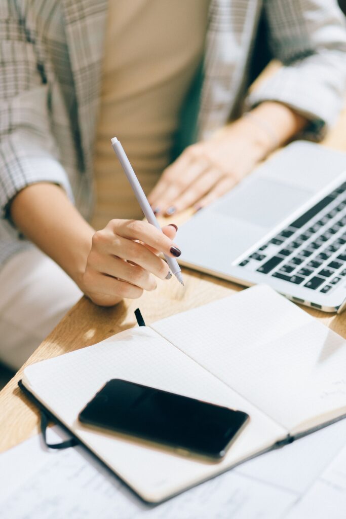 pexels photo 5186375 5186375 Close-up of a person writing notes on a notebook next to a laptop and smartphone on a workspace.