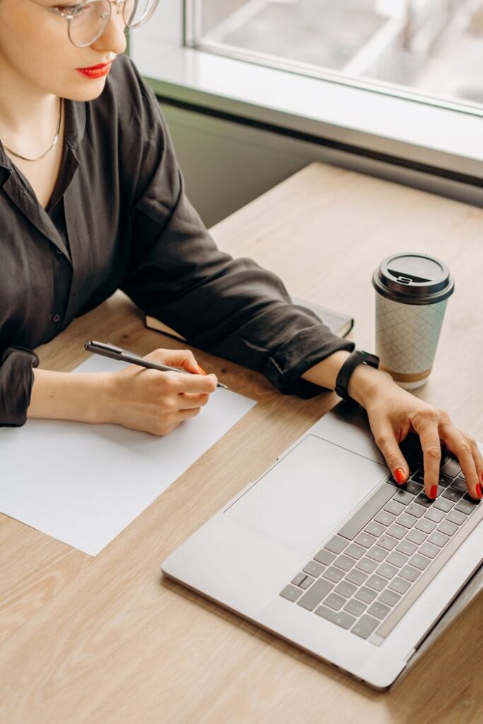 pexels photo 8297056 Businesswoman writing notes while working on a laptop at a wooden desk with a coffee cup nearby.