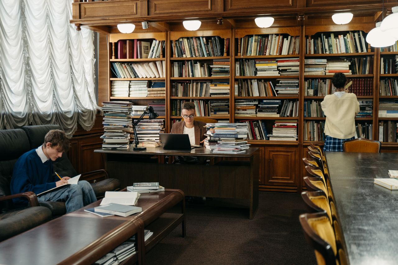 about-me Students studying in a spacious library, surrounded by wooden shelves filled with books.